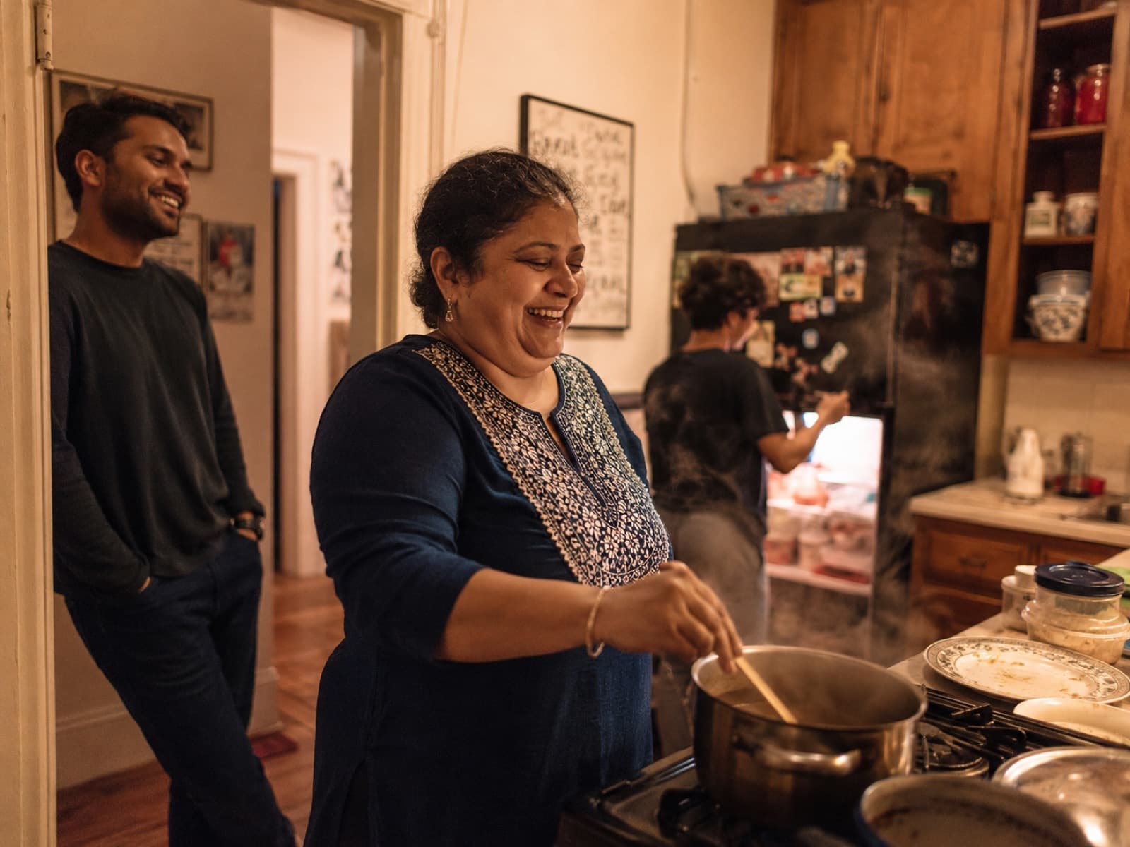 A woman laughing while cooking at the stove as her family gathers in a warm, lived-in home kitchen.
